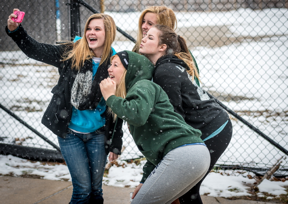 Vier high school studentes in winterkleding nemen samen een selfie buiten op een besneeuwde dag, staand voor een hek van gaas. Ze hebben duidelijk plezier