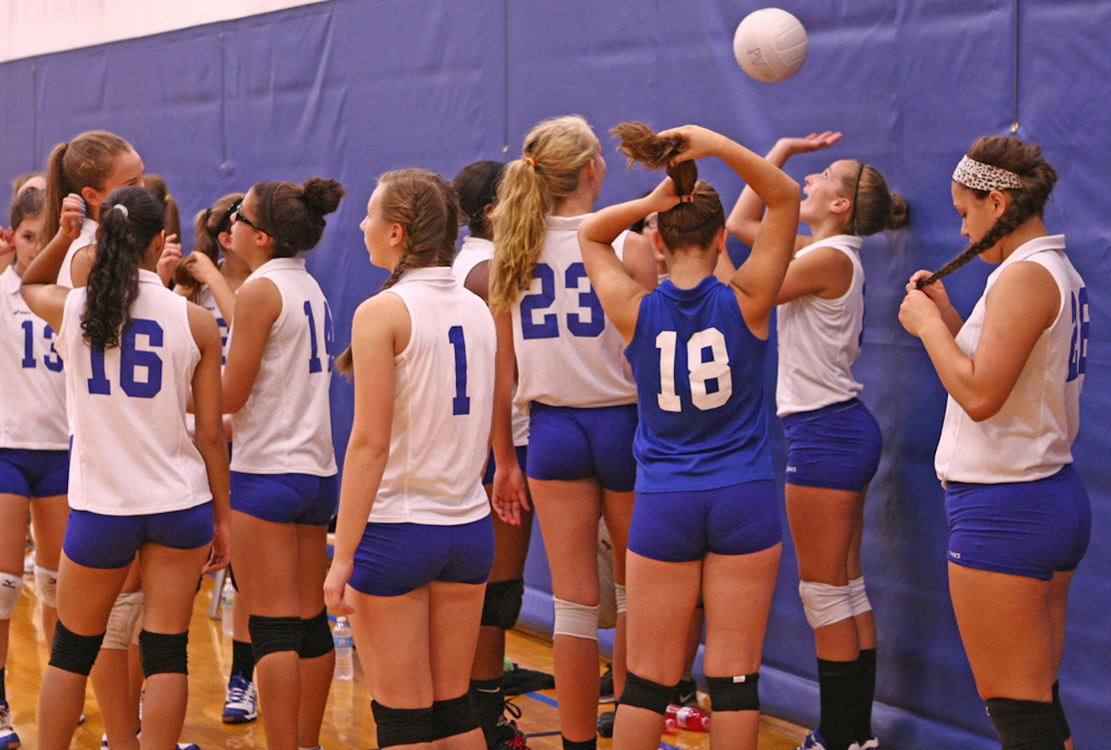 Een groep vrouwelijke volleybalspelers in blauw-witte tenues staat samen in een gymzaal. Een van de spelers gooit een volleybal over zijn hoofd.