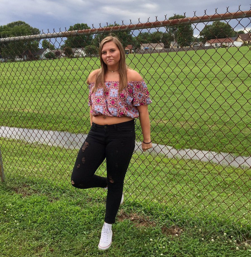 Een high school studente staat voor een hek van gaas dat een schoolterrein omzoomt. Ze heeft kortgeleden straf op haar billen gekregen terwijl ze een yogabroek aanhad en daar kwamen de klappen hard door heen. Ze draagt nu een off-shoulder top met bloemenprint, een zwarte spijkerbroek met scheuren en witte sneakers.