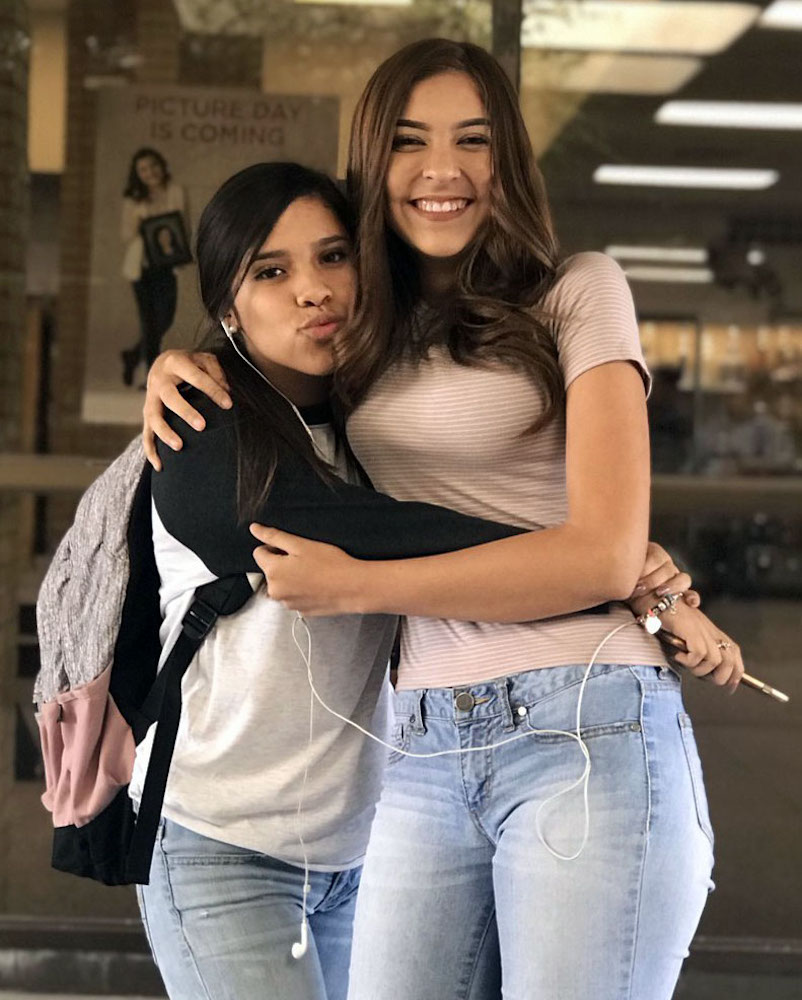Twee studentes poseren en glimlachen terwijl ze elkaar omhelzen in een gang. De een houdt een potlood vast en beiden hebben oordopjes in. Het grootste meisje heeft kort daarvoor voor de eerste keer van haar leven straf op haar billen gekregen. Op de achtergrond is een poster met de tekst