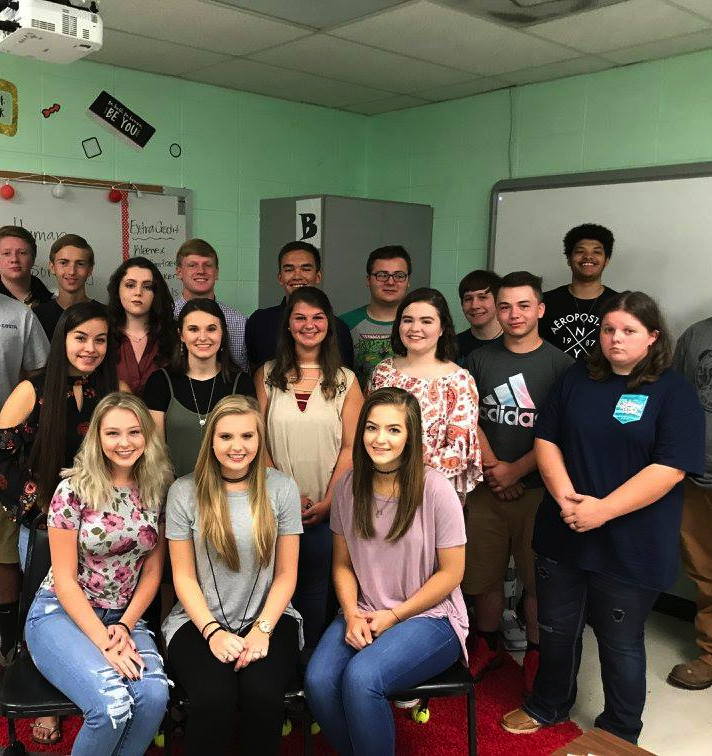 Een groep van zeventien High School studenten poseert voor een groepsfoto in een klaslokaal. Sommigen zitten vooraan en anderen staan achteraan.