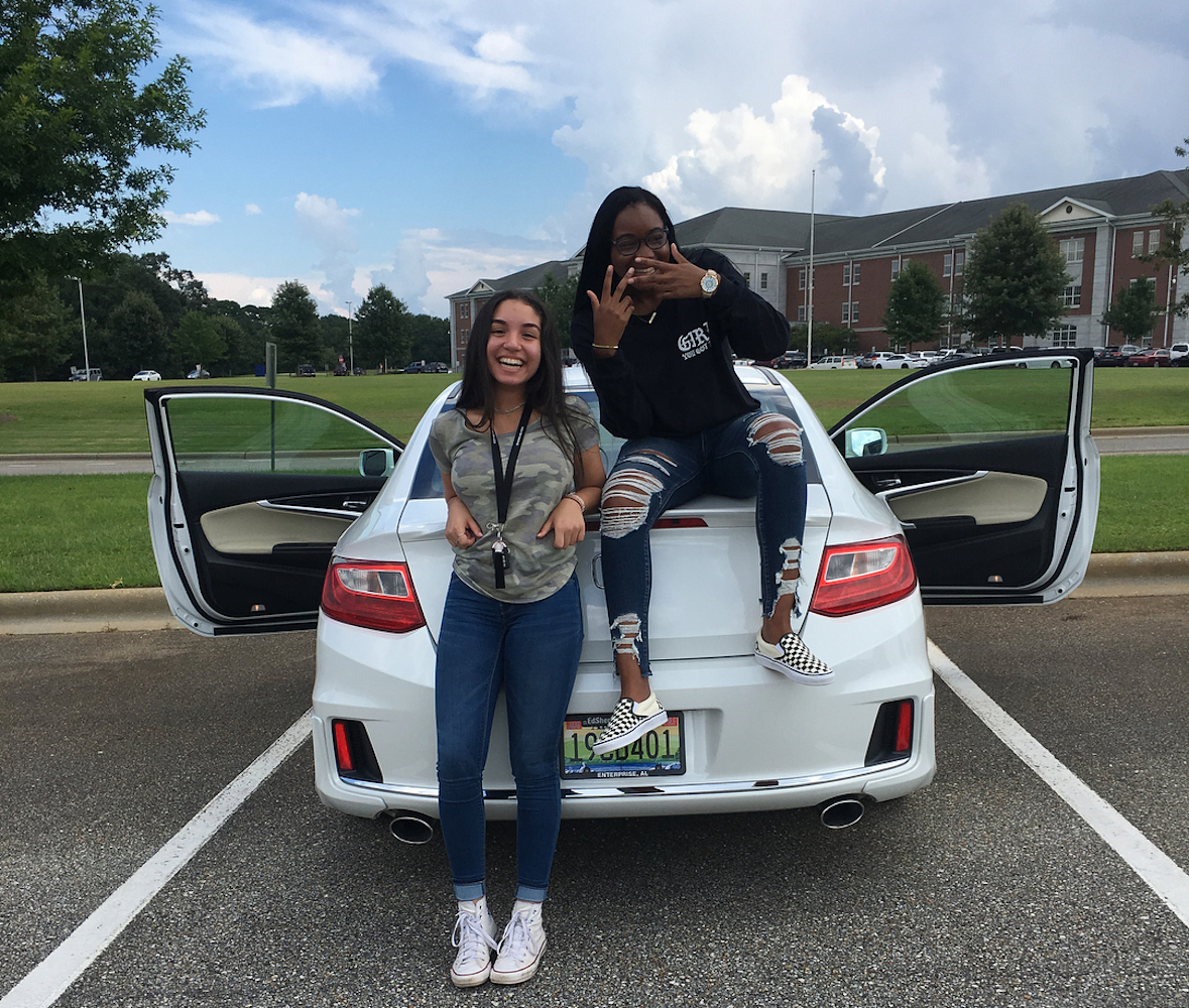 Twee studentes poseren voor een witte auto met open deuren op een parkeerplaats. De één staat en de ander zit op de kofferbak van de auto. Op de achtergrond is het schoolgebouw zichtbaar.
