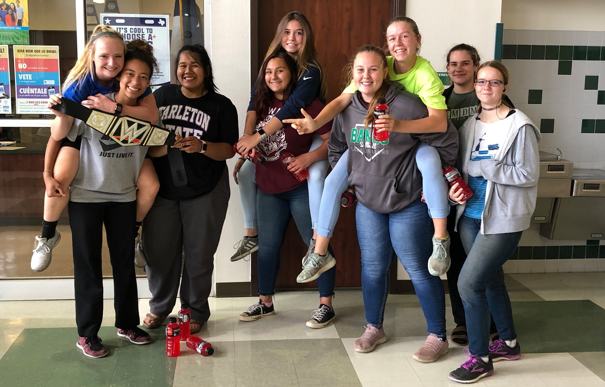 Een groep van acht lachende high school studentes poseert binnen. Twee van hen dragen kampioensgordels en meerdere hebben waterflessen. Twee vrouwen rijden op de rug van de vrouwen.