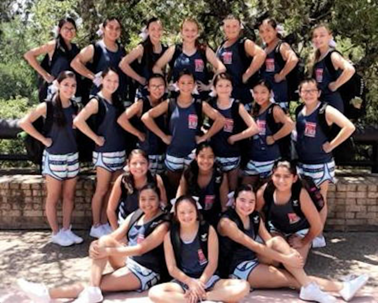 Een groep van achttien high school cheerleaders in bijpassende sportkleding poseert buiten. Zestien vrouwen staan en twee zitten met hun benen gekruist op de voorgrond, en ze lachen allemaal naar de camera.