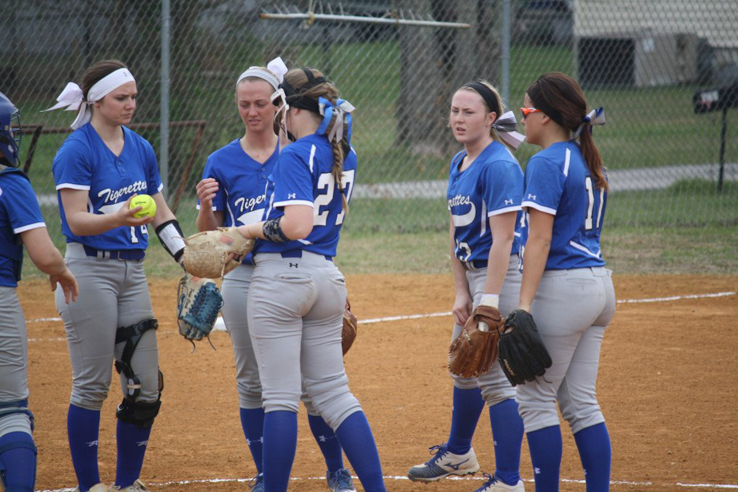 Een groepje softbalspelers in blauwe uniformen staat samen op het veld, met handschoenen en een bal in de hand, en voert een gesprek bij de werpheuvel.