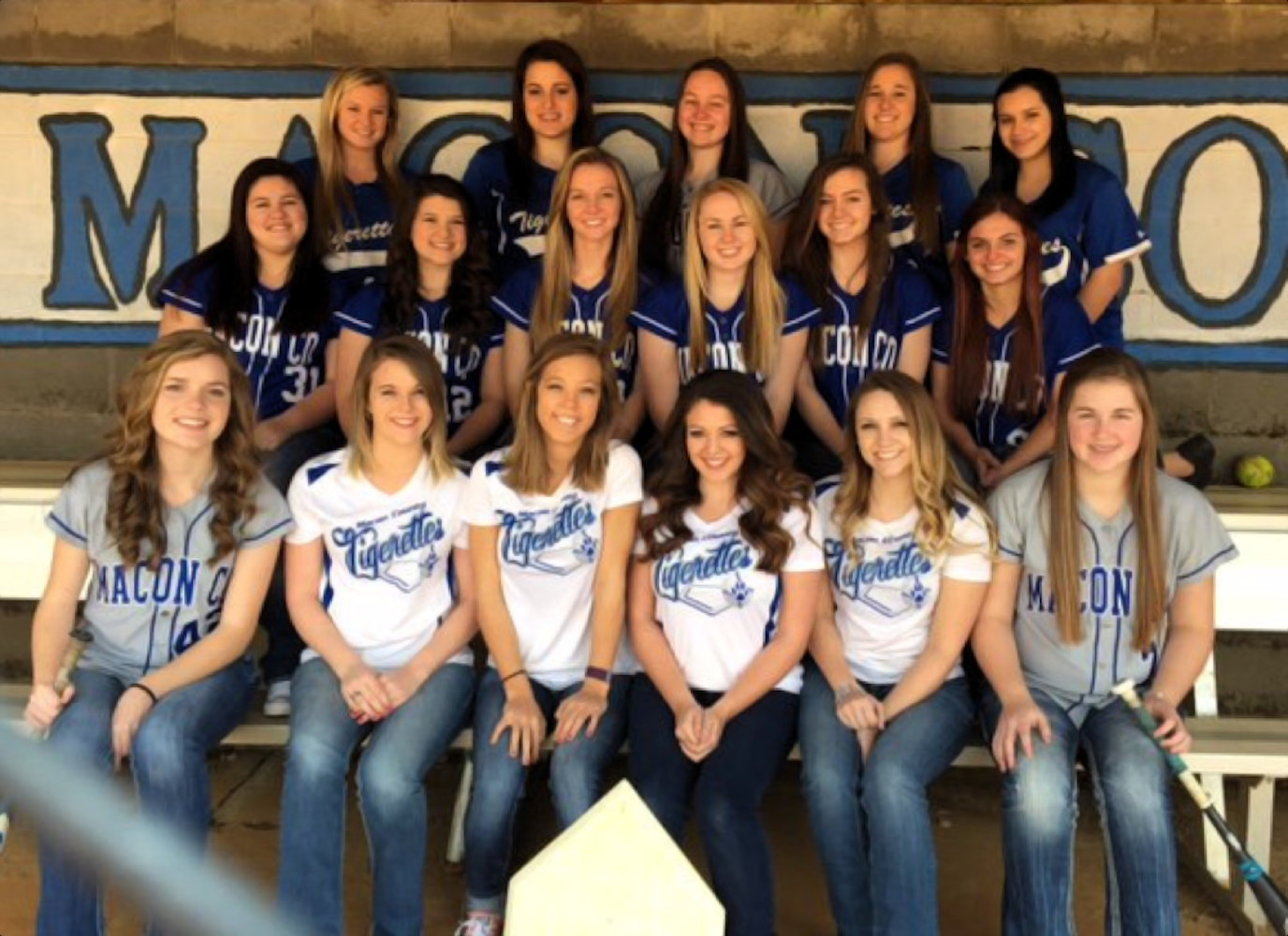 Een groep high school studentes van de Macon HIgh School in softbaluniformen poseert voor een teamfoto op een bankje voor een gebouw met een