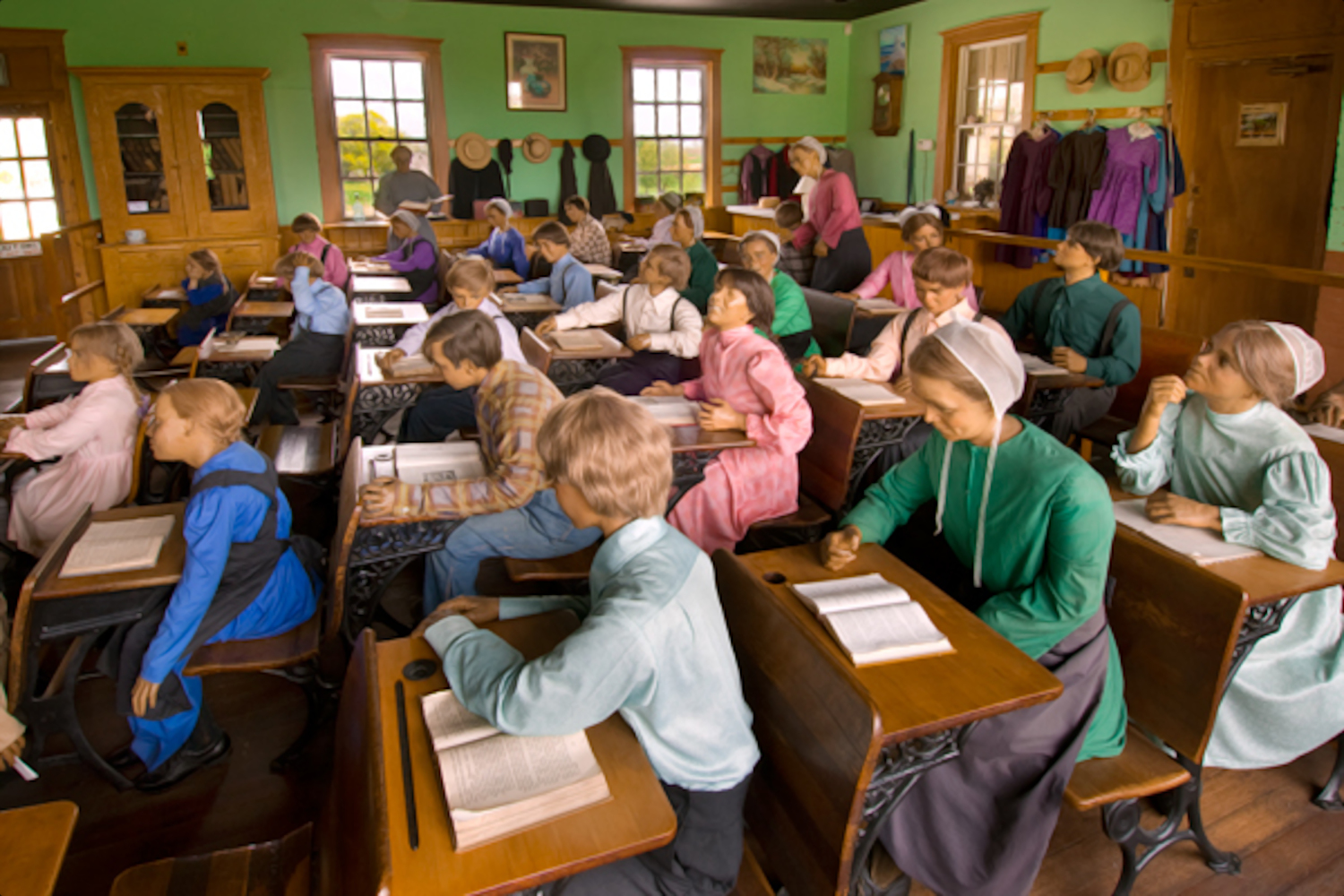 Kinderen in traditionele Amsih kleding zitten aan houten tafels in een klaslokaal met groene wanden en een leraar achterin. Boeken en schrijfgerei liggen op de tafels.