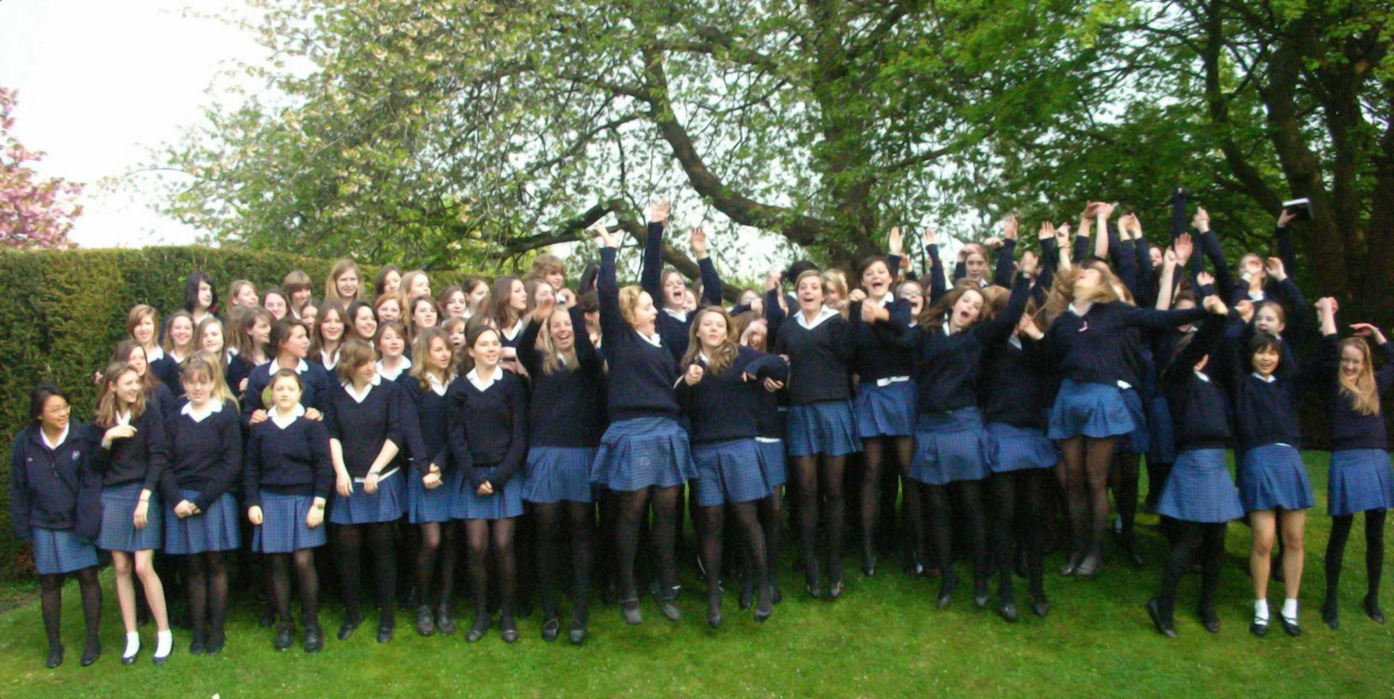 Een grote groep vrouwelijke studenten van een Christelijke privé school in blauwe uniformen staat buiten op een grasveld te springen voor een foto, met bomen en heggen op de achtergrond. Bij toelating op deze school moeten ouders en studenten dat ze akkoord zijn met billenkoek als disciplinaire maatregel.