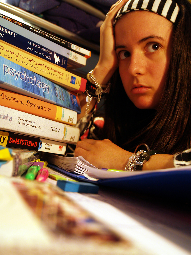 Een jongere zit aan een bureau met zijn hoofd rustend op zijn hand, omringd door een stapel psychologieboeken, schriften en schrijfbenodigdheden, op zoek naar overzicht en ondersteuning in zijn studie.