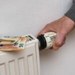 Een hand stelt een radiatorthermostaat af met eurobankbiljetten bovenop de radiator.