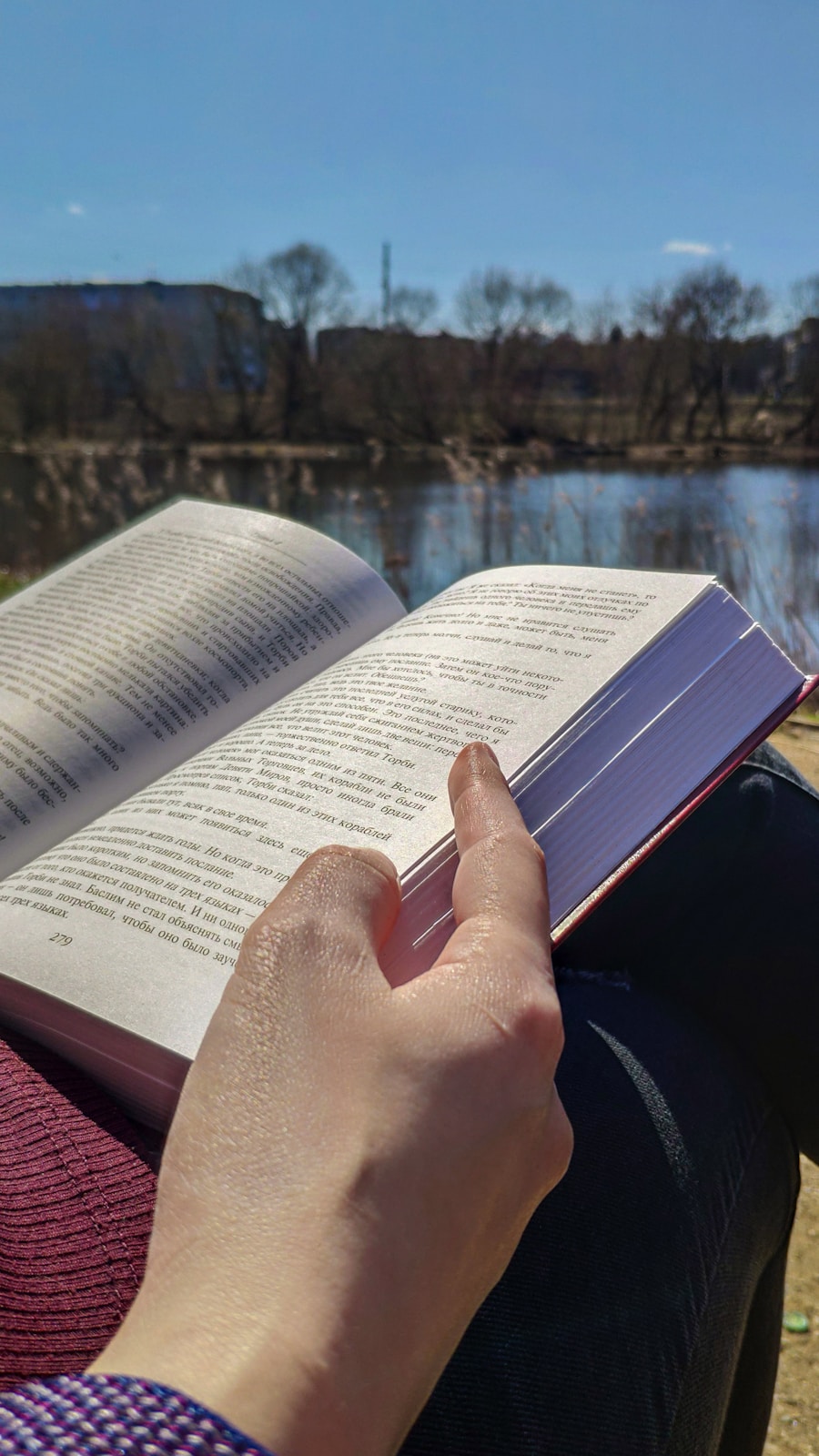 Een persoon leest een open boek buiten bij een meer op een zonnige dag, met bomen en gebouwen zichtbaar op de achtergrond.