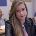 Een student in een zwart leren jack zit aan een bureau in een klaslokaal, kijkt geconcentreerd voor zich uit en belichaamt zelfstandig werken, terwijl twee andere studenten onscherp op de achtergrond te zien zijn.