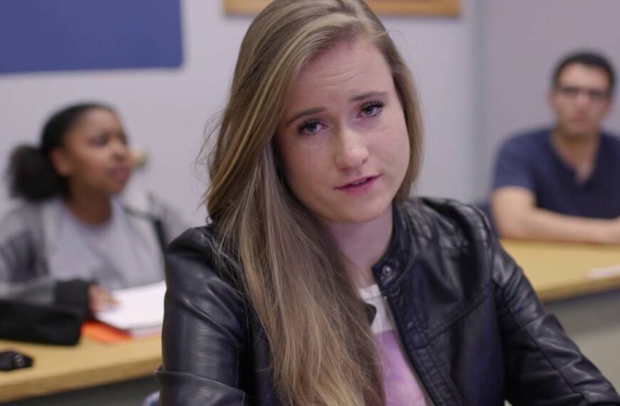 Een student in een zwart leren jack zit aan een bureau in een klaslokaal, kijkt geconcentreerd voor zich uit en belichaamt zelfstandig werken, terwijl twee andere studenten onscherp op de achtergrond te zien zijn.