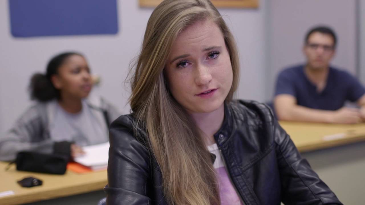 Een student in een zwart leren jack zit aan een bureau in een klaslokaal, kijkt geconcentreerd voor zich uit en belichaamt zelfstandig werken, terwijl twee andere studenten onscherp op de achtergrond te zien zijn.