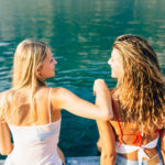 Twee vrouwen in zwemkleding zitten op een steiger aan helder water, de een met haar arm om de ander heen, allebei kijken ze elkaar aan en glimlachen.