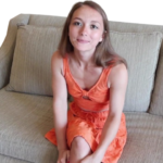 Een vrouw in een oranje jurk zit op een beige bank, kijkt met een lichte glimlach naar de camera en vangt een moment van aansluiting vinden temidden van eenzaamheid.