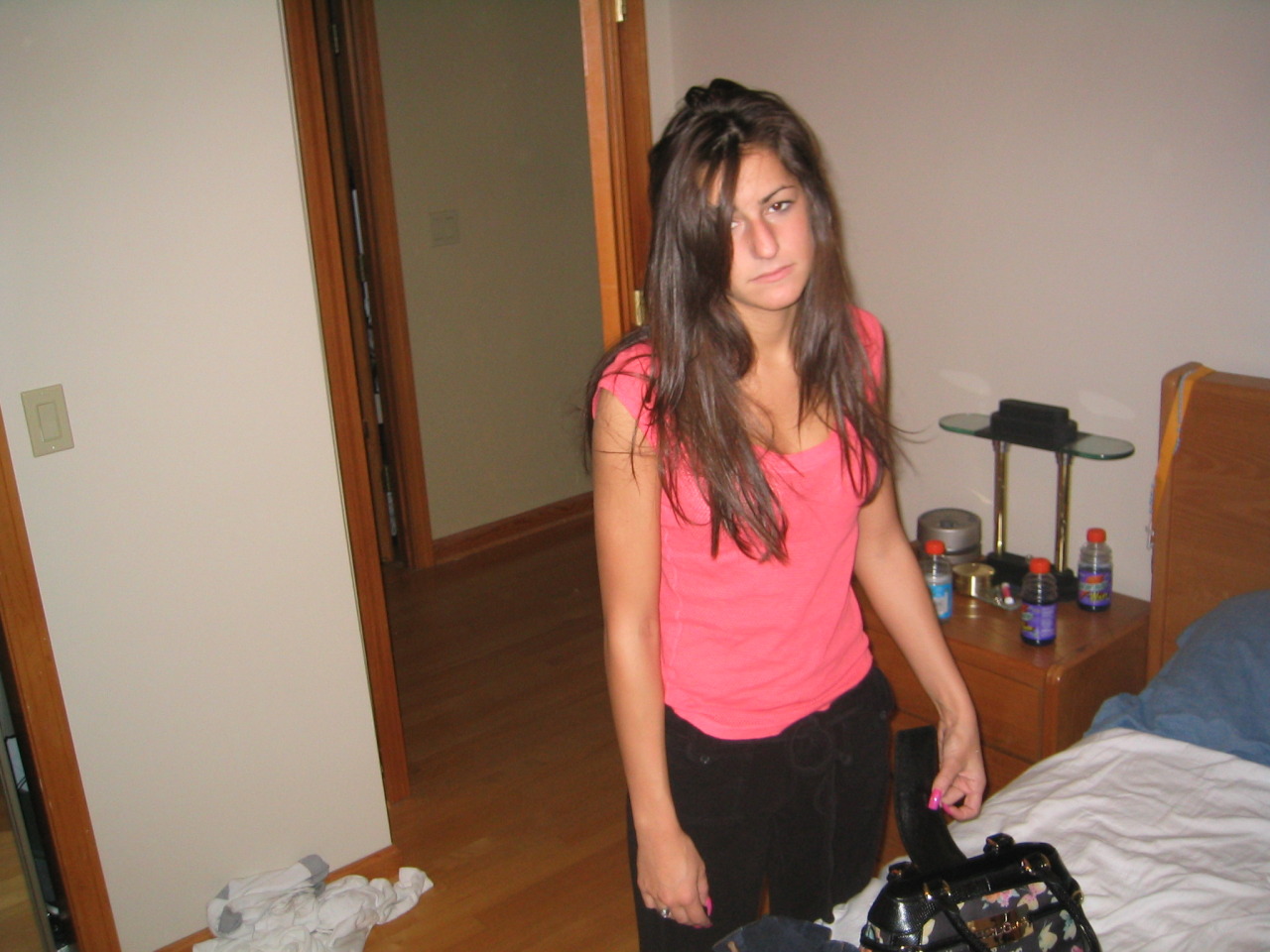 Een vrouw met lang bruin haar, in een roze shirt en zwarte broek, staat in een slaapkamer naast een bed met spullen erop en kijkt naar de camera met een neutrale uitdrukking die wijst op eenzaamheid of moeite om aansluiting te vinden.
