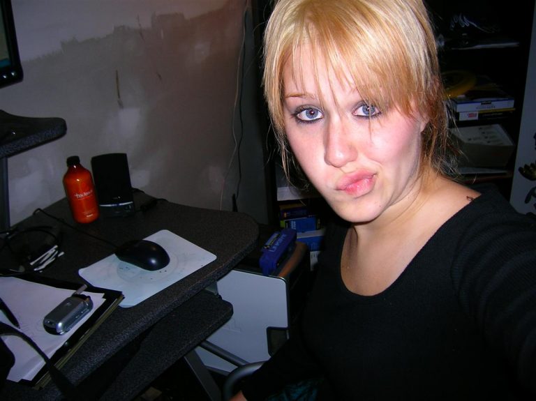 Student met blonde haren maakt een lichte grijns terwijl ze een selfie neemt aan haar rommelige bureau, omringd door computer, muis en diverse spullen-perfect voor zelfstandig werken en zelfstandig denken.