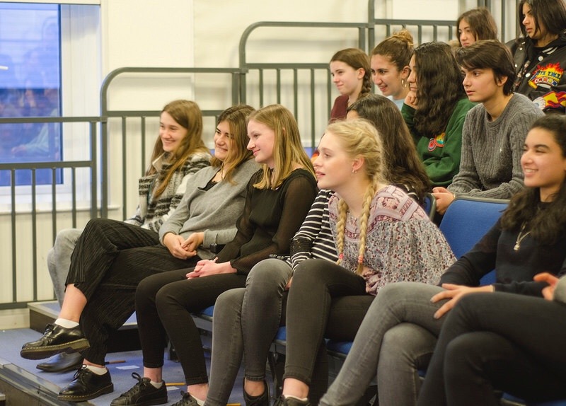Een groep studenten zit op blauwe tribunes in een klaslokaal of aula, aandachtig te kijken naar iets dat niet te zien is, klaar om zelfstandig denken en zelfstandig werken te oefenen.