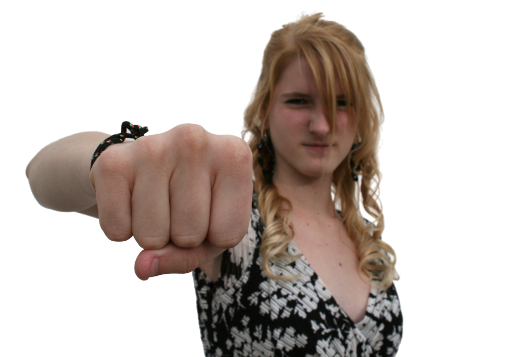 Een jonge vrouw met lang blond haar steekt haar vuist uit naar de camera, draagt een jurk met patroon en een zwarte armband, tegen een neutrale witte achtergrond - een weergave van de zelfdruk die velen voelen als ze thuis moeten presteren.