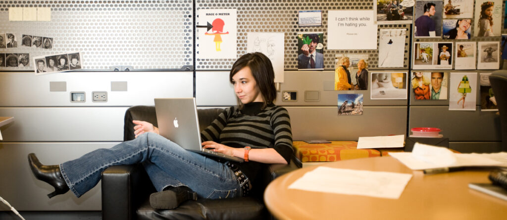 Een vrouw zit op een zwarte stoel met haar benen gekruist, ze werkt op een laptop in een kantoor vol foto's en notities, omringd door prestatiedruk terwijl ze probeert uit te blinken in haar taken.