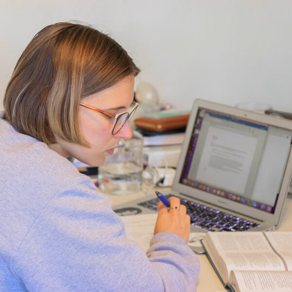 Een student met een bril zit aan een bureau en maakt aantekeningen terwijl hij een open boek en een document op een MacBook laptop raadpleegt.