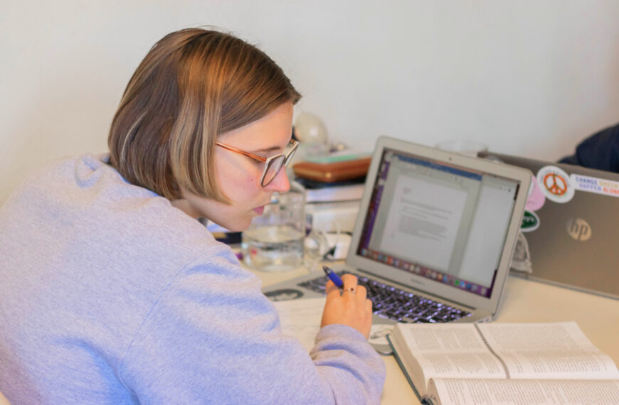 Een student met een bril zit aan een bureau en maakt aantekeningen terwijl hij een open boek en een document op een MacBook laptop raadpleegt.