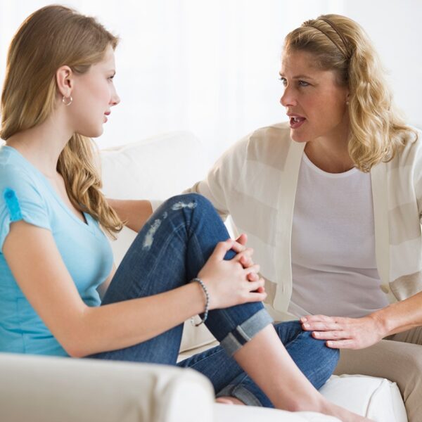 Een vrouw en een tienermeisje zitten op een bank tegenover elkaar en hebben een serieus gesprek over presteren en omgaan met druk thuis in een lichte woonkamer.