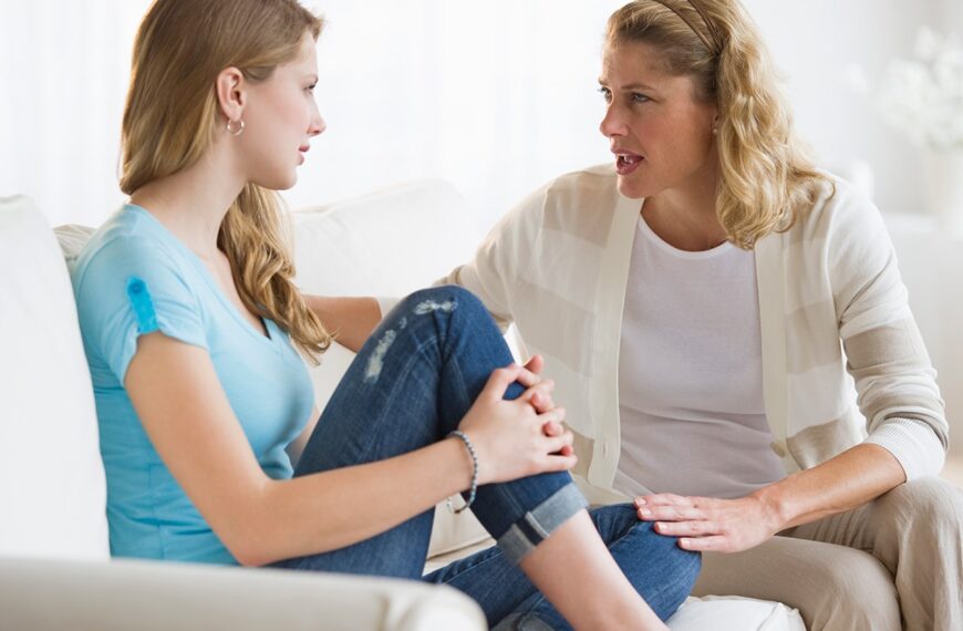 Een vrouw en een tienermeisje zitten op een bank tegenover elkaar en hebben een serieus gesprek over presteren en omgaan met druk thuis in een lichte woonkamer.