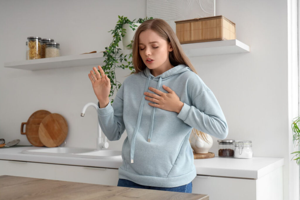 Een jonge vrouw met een blauwe capuchon staat in een keuken, houdt met één hand haar borst vast en steekt de andere hand op, terwijl ze ongemak of kortademigheid lijkt te ervaren.