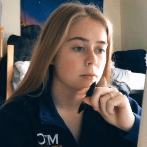 Een jonge vrouw met lang blond haar zit aan een bureau, houdt een pen onder haar kin en kijkt met een nadenkende blik naar het computerscherm. Ze vindt het moeilijk om zich op haar studie te concentreren.