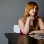 Een vrouw met lang rood haar zit aan een tafel met verschillende papieren bankbiljetten voor haar uitgespreid en kijkt met een neutrale uitdrukking opzij.