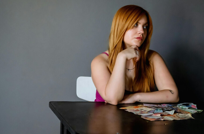 Een vrouw met lang rood haar zit aan een tafel met verschillende papieren bankbiljetten voor haar uitgespreid en kijkt met een neutrale uitdrukking opzij.