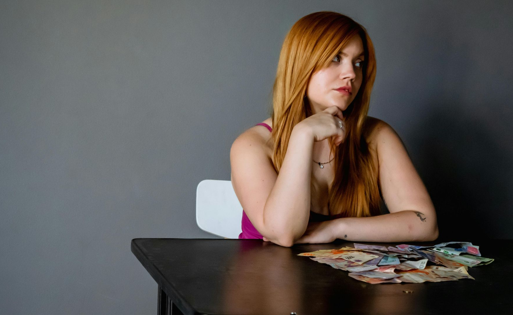 Een vrouw met lang rood haar zit aan een tafel met verschillende papieren bankbiljetten voor haar uitgespreid en kijkt met een neutrale uitdrukking opzij.