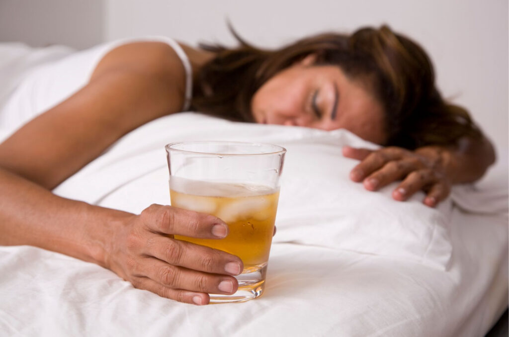 Een vrouw ligt in bed met een glas ijskoude amberkleurige vloeistof in haar hand, ze lijkt te slapen of te rusten.
