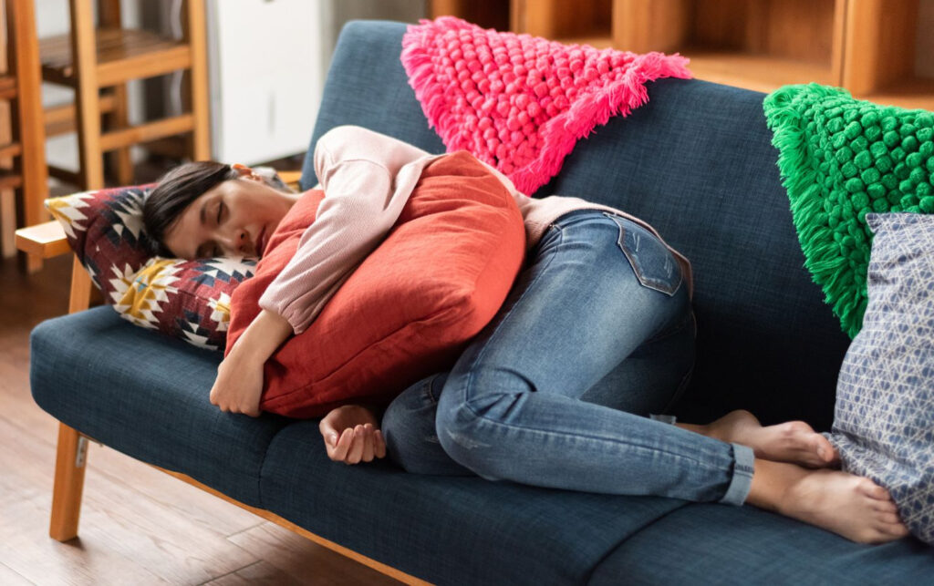 Een persoon in jeans en een roze topje ligt op een blauwe sofa, omhelst een oranje kussen en rust uit met gesloten ogen. Achter hen liggen felgekleurde kussens.