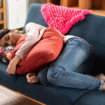 Een persoon in jeans en een roze topje ligt op een blauwe sofa, omhelst een oranje kussen en rust uit met gesloten ogen. Achter hen liggen felgekleurde kussens.