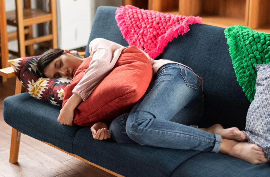 Een persoon in jeans en een roze topje ligt op een blauwe sofa, omhelst een oranje kussen en rust uit met gesloten ogen. Achter hen liggen felgekleurde kussens.
