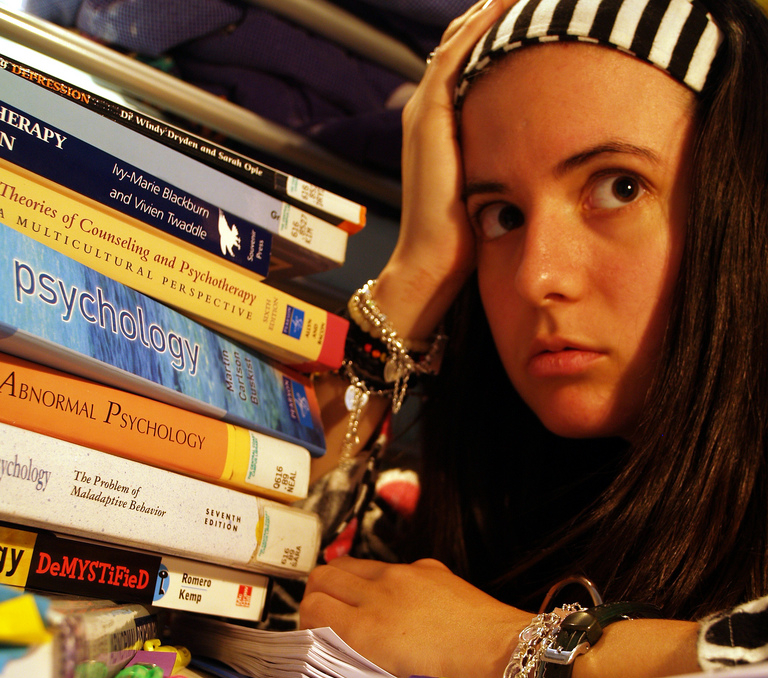 Een jongere met een gestreepte hoofdband kijkt gestrest terwijl hij naast een hoge stapel psychologie- en therapieboeken zit.