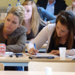 Studentes zitten in een collegezaak; de ene luistert aandachtig terwijl de andere aantekeningen maakt. Andere mensen zitten op de achtergrond. Flesjes water en papieren bekertjes staan op de bureaus. Eén studente kijkt verbijsterd.