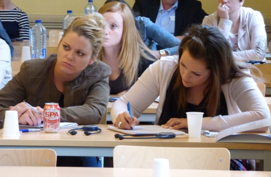 Studentes zitten in een collegezaak; de ene luistert aandachtig terwijl de andere aantekeningen maakt. Andere mensen zitten op de achtergrond. Flesjes water en papieren bekertjes staan op de bureaus. Eén studente kijkt verbijsterd.