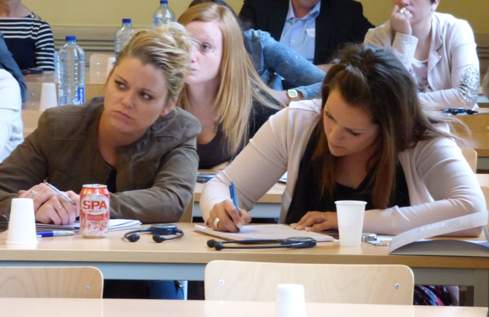 Studentes zitten in een collegezaak; de ene luistert aandachtig terwijl de andere aantekeningen maakt. Andere mensen zitten op de achtergrond. Flesjes water en papieren bekertjes staan op de bureaus. Eén studente kijkt verbijsterd.